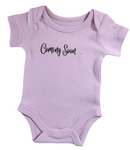 Pink baby onesie with 'Coming Soon' text on a wooden surface with blue flowers in the background